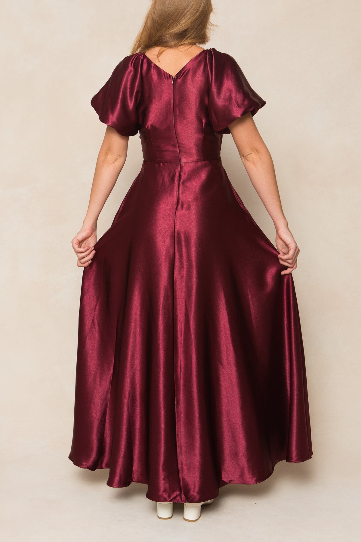 A woman faces away in the Ivy City Co Tessie Dress in Wine Satin, featuring short puff sleeves and a flared, floor-length skirt. She holds the sides of this elegant fall bridesmaid dress while white shoes peek out beneath the hem.