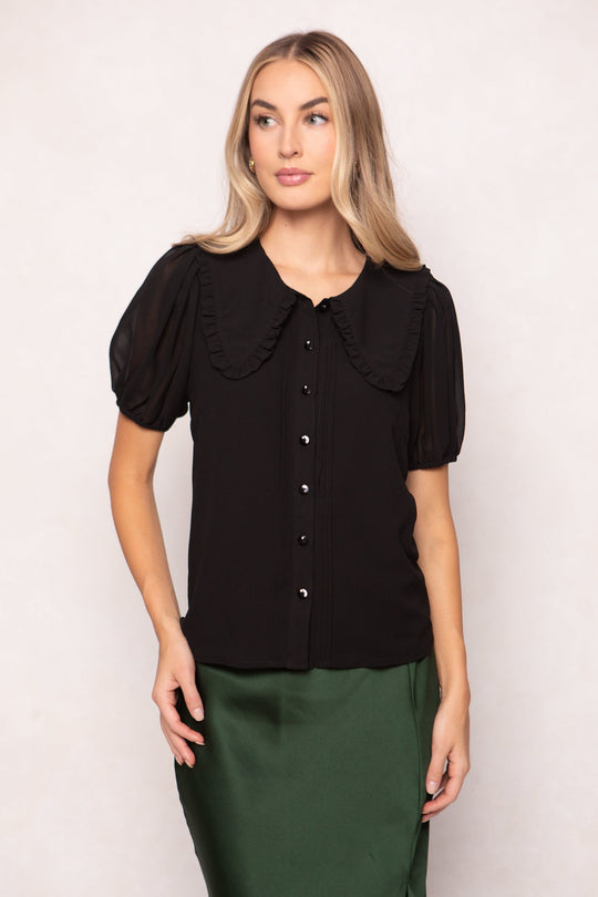 A woman wearing the Ivy City Co Andrea Blouse in Black Chiffon, featuring puff sleeves and a ruffled collar, paired with a green satin skirt, stands against a plain, light background.
