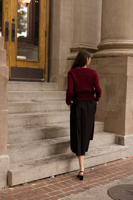 Wearing the Theo Sweater in Burgundy by Ivy City Co, a woman with her hands in her pockets walks up stone steps toward large wooden doors, flanked by tall columns, pairing her cozy fall knit with a black skirt.
