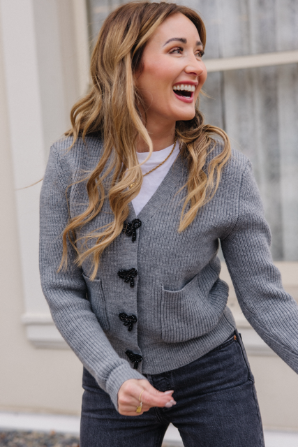 A smiling woman stands outside in front of a window, laughing as she wears the Ivy City Co Theo Sweater in Gray over a white shirt and dark jeans, radiating effortless style.
