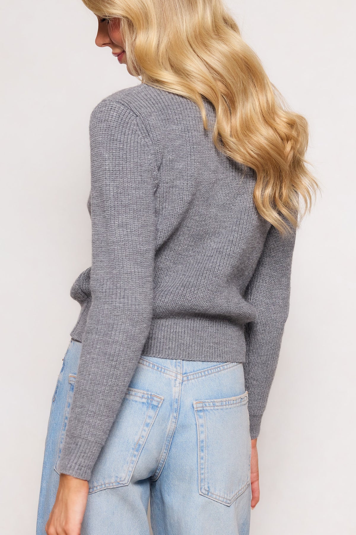 A person with long, wavy blonde hair models the Ivy City Co Theo Sweater in Gray and light blue jeans, standing slightly turned against a simple, light background.