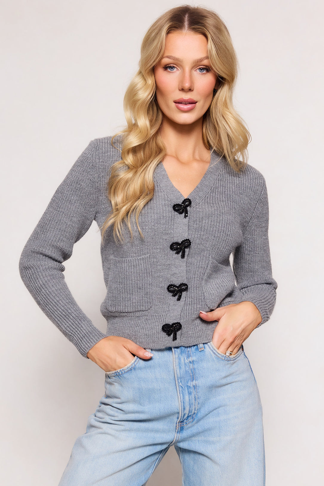 A woman with long blonde hair wears the Ivy City Co Theo Sweater in Gray paired with light blue high-waisted jeans, standing with one hand in her pocket against a light background.