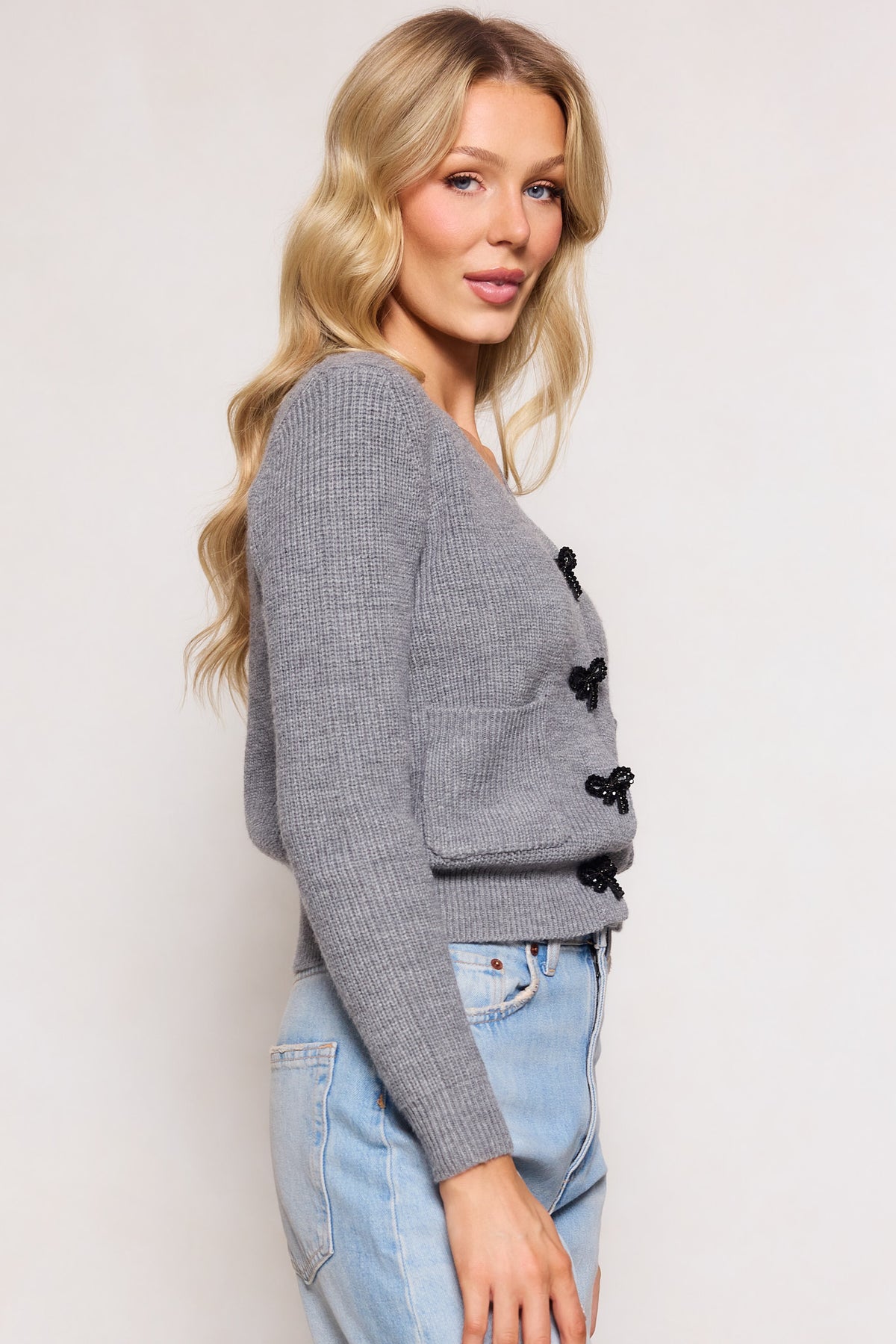 A woman with long blonde hair models the Ivy City Co Theo Sweater in Gray—a light gray, ribbed cardigan with black decorative buttons and a structured v-neckline—paired with light blue jeans against a plain background.