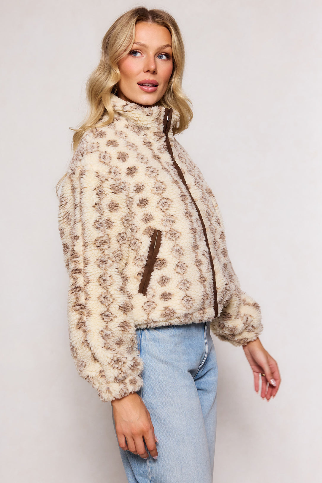 A woman wears the Ivy City Co Theodore Jacket in Tan & White, featuring brown trim and structured seams, paired with light blue jeans. She stands sideways against a plain background, looking slightly toward the camera.