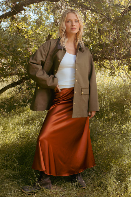 A woman stands on grass under leafy trees, wearing an olive jacket, white top, and the Ivy City Co Tiff Maxi Skirt in Copper with brown boots. She looks at the camera with one hand on her hip.