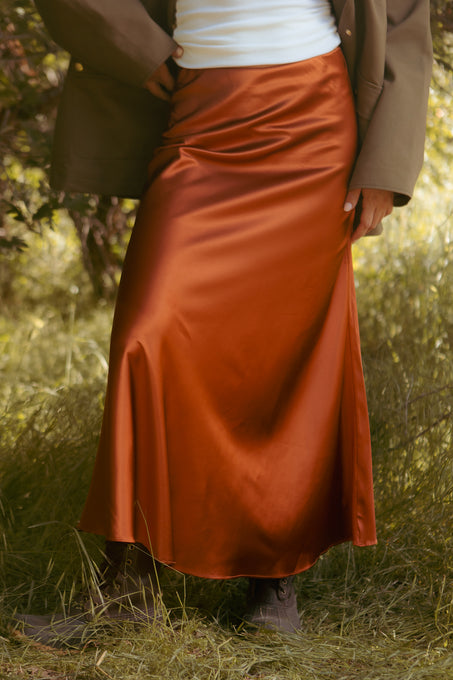 A person stands on grass wearing the Ivy City Co Tiff Maxi Skirt in Copper with a white top, olive green jacket, and brown boots. Their face is not visible, emphasizing the elegant satin skirt and chic look.