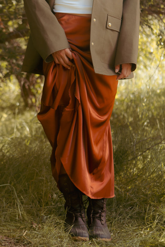 Standing outdoors on grass, a person in dark boots, a white top, and an olive green jacket holds up the Ivy City Co Tiff Maxi Skirt in Copper; their face is not visible.