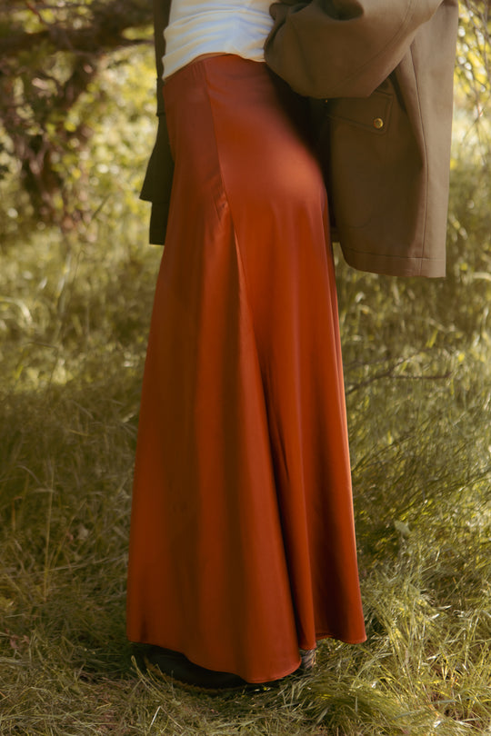 Outdoors on grass, a person wears the Ivy City Co Tiff Maxi Skirt in Copper with a white top and an olive green jacket over their shoulders. Their upper body and face are not visible.