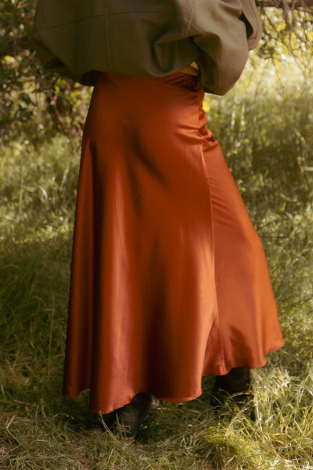 Outdoors on grass, a person wears the Ivy City Co Tiff Maxi Skirt in Copper and a loose olive green jacket, with their face out of frame.