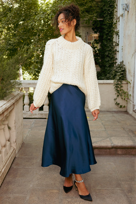Woman wearing a white sweater and blue skirt standing outdoors.