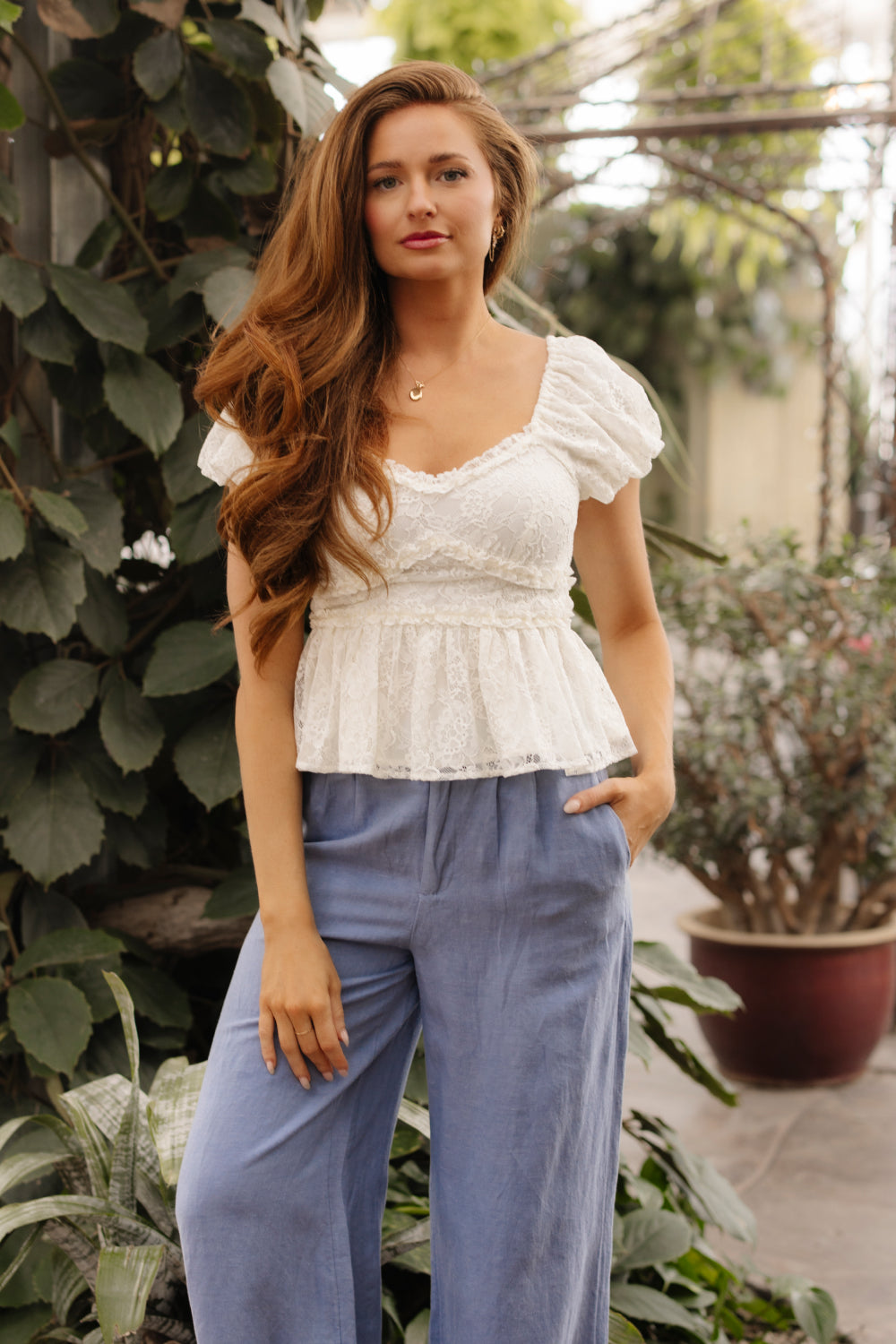 A woman with long, wavy brown hair wears the Ivy City Co Torino Top in Ivory Lace with ruffle details and puffed sleeves, paired with light blue wide-leg pants, as she stands surrounded by lush greenery.