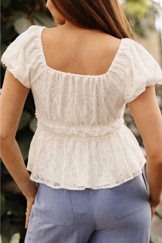 A woman with long brown hair is seen from behind wearing the Ivy City Co Torino Top in Ivory Lace, featuring ruffle details and a square neckline, paired with light blue pants. Green foliage appears blurred in the background.