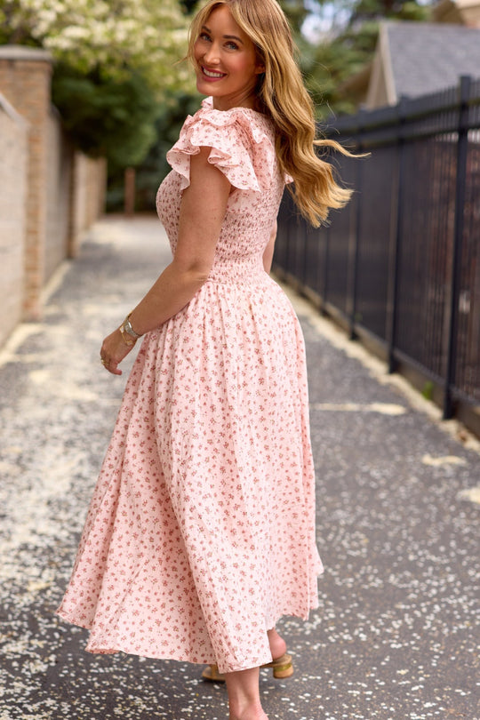Wearing the Hattie Dress in Madeline Pink by Ivy City Co, a smiling woman with ruffled sleeves walks down a tree-lined path, looking back over her shoulder.