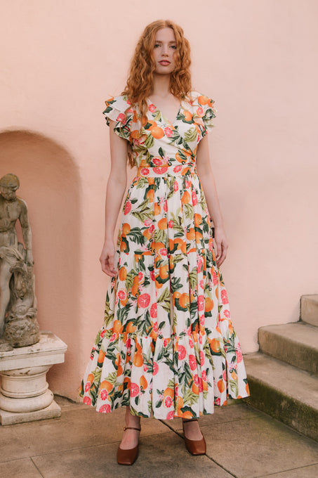 A woman with long red hair wears the Ivy City Co Trixie Dress in Citrus, a vibrant fruit-printed cotton maxi with ruffles and brown heels, standing on stone steps by a classical statue against a pale pink wall.