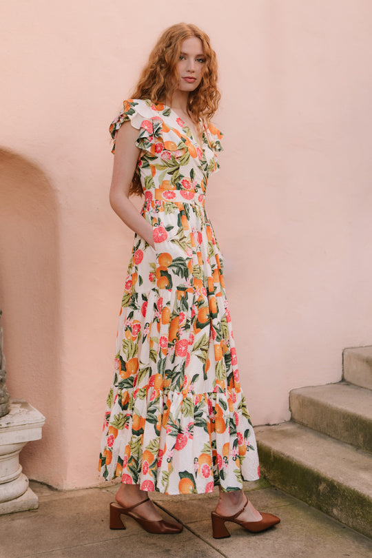 A woman with long, wavy red hair wears the Ivy City Co Trixie Dress in Citrus, a white cotton maxi floral dress with ruffled sleeves, and brown block-heeled shoes, standing beside a pink wall and stone steps.