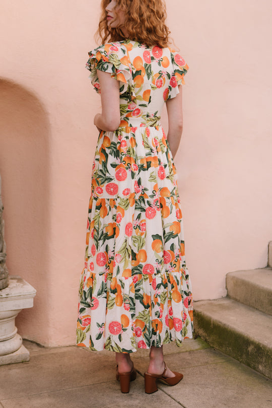 A woman with curly red hair wears the Ivy City Co Trixie Dress in Citrus, a maxi-length cotton dress with a colorful fruit pattern, and brown block-heeled shoes as she stands on a stone path near steps and a pale peach wall.