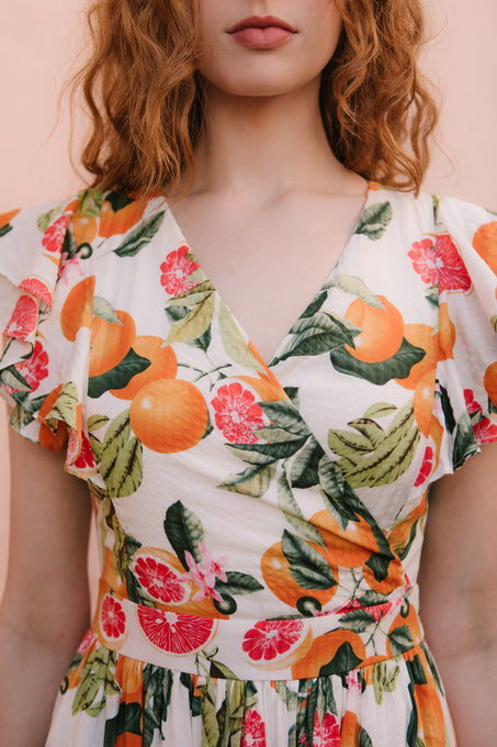 A woman with wavy red hair models the Ivy City Co Trixie Dress in Citrus, featuring a bold pattern of oranges, green leaves, and pink flowers. The image is cropped from shoulders to waist to showcase the dress’s vibrant design.