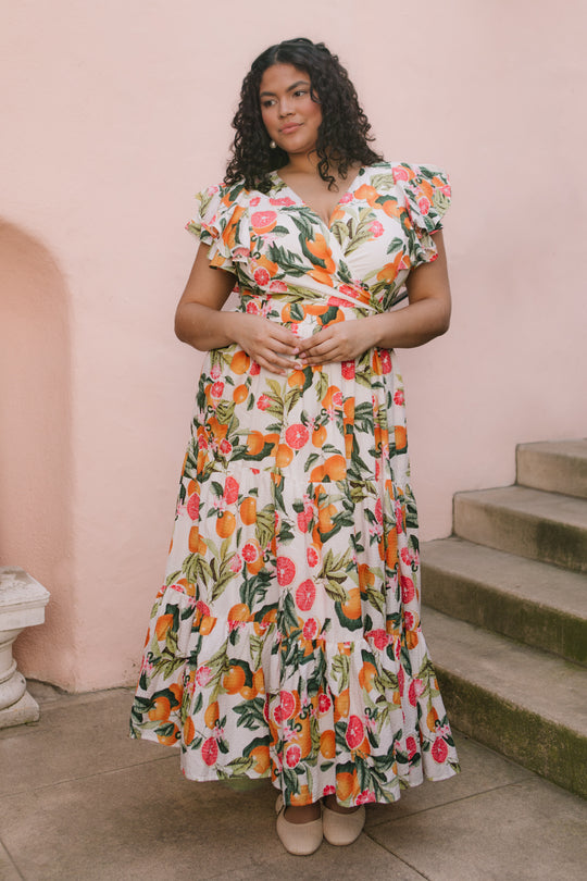 A woman with curly hair, wearing the Ivy City Co Trixie Dress in Citrus—a colorful, maxi cotton dress with ruffles—stands by concrete steps against a pink wall, clasping her hands and gazing to the side.