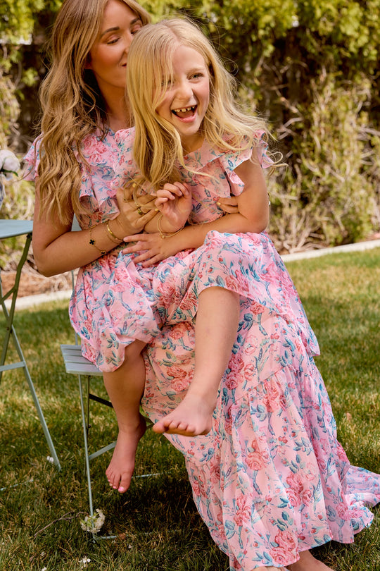 A woman and young girl sit together in a garden, both smiling and relaxed in matching Ivy City Co Trixie Dresses in Pink Sketched Flower Chiffon—perfect stylish outfits for a summer wedding.