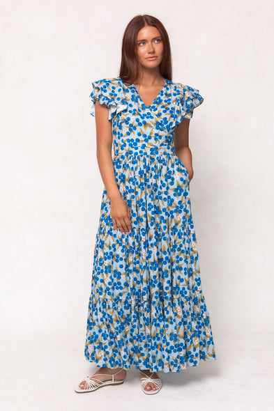 A woman with long brown hair wears the Ivy City Co Trixie Dress in Blueberry—a cotton maxi dress with ruffled short sleeves and pockets—while standing against a plain white background in white sandals.