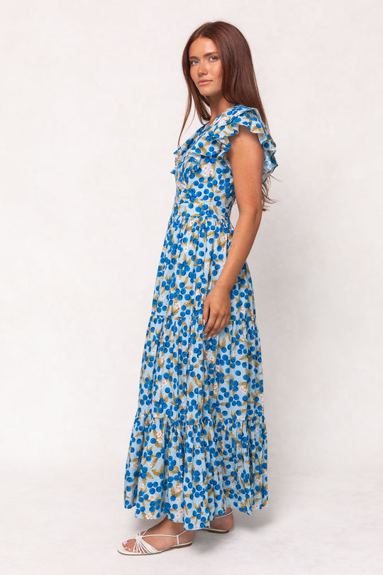 A woman with long brown hair wears the Ivy City Co Trixie Dress in Blueberry—a sleeveless, ankle-length cotton maxi dress with ruffled straps—and white sandals, standing sideways against a plain white background.