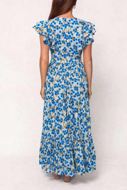 A woman faces away, wearing the Ivy City Co Trixie Dress in Blueberry—a long cotton maxi dress with short ruffled sleeves, a fitted waist, and a tiered skirt featuring blue, yellow, and green floral patterns on white.