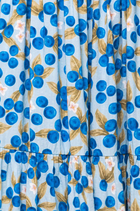A close-up of the Trixie Dress in Blueberry by Ivy City Co shows gathered fabric with a vibrant blue background, patterned with clusters of blue berries, green leaves, and small white flowers.