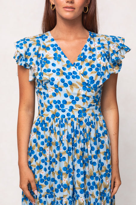 Woman wearing the Ivy City Co Trixie Dress in Blueberry—a blue and white floral-patterned cotton maxi dress with ruffled short sleeves and a V-neckline—standing against a plain light background.