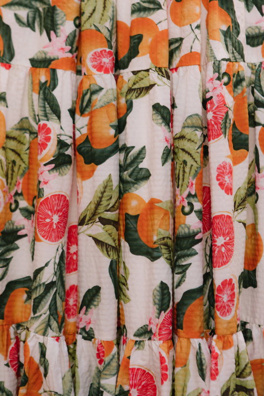 A close-up of the Trixie Dress in Citrus by Ivy City Co shows a white, textured cotton fabric with oranges, grapefruits, green leaves, and pink flowers. The dress features soft gathers and vibrant citrus patterns.