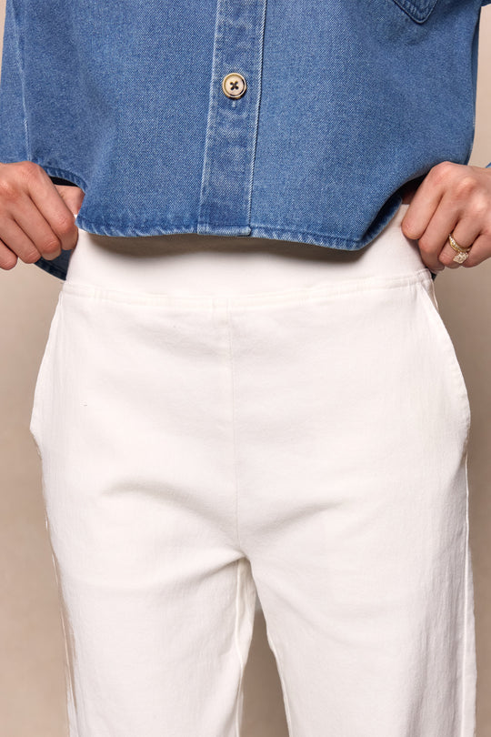 A person wears Ivy City Co’s Tyler Pants in Cream, styled with a blue denim shirt tucked in and hands resting on the waistband. Only the torso and upper legs are visible.