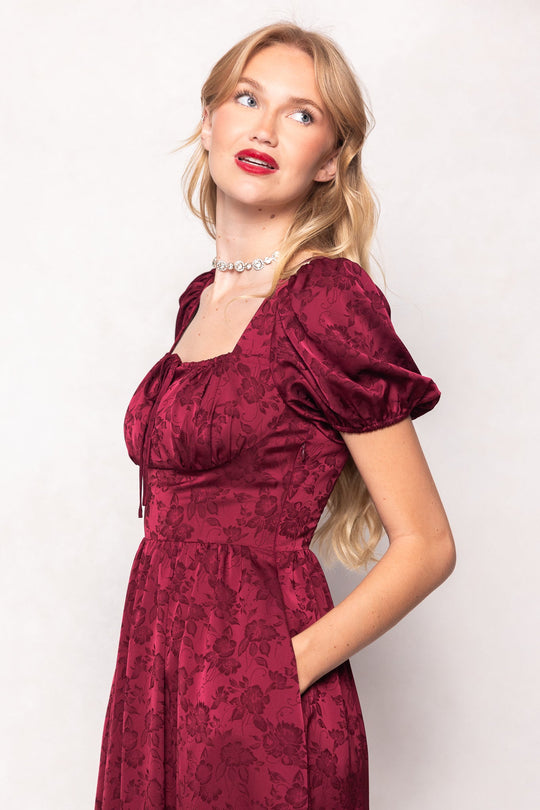 A woman with long blonde hair, red lipstick, and a pearl necklace stands against a light background, looking up and to her left while wearing the Ivy City Co Amira Dress in Red with a floral pattern and puff sleeves.