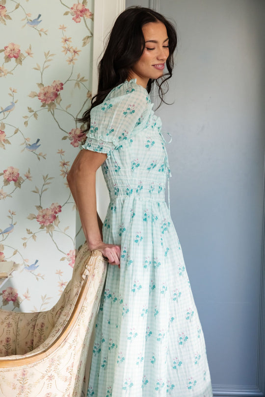 A woman wearing the Jessie Dress in Bloom Gingham by Ivy City Co stands beside a cream-colored chair with floral upholstery, against a wall adorned with pink flower wallpaper, looking down and smiling gently.