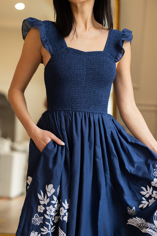 A woman wearing the Finch Dress in Blue by Ivy City Co—a deep blue cotton dress featuring ruffled straps and delicate white embroidery near the hem—stands indoors, one hand holding the dress, the other tucked in a pocket.