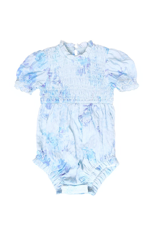 Introducing the Baby Flynn Romper in Aquarelle Fleur by Ivy City Co—a light blue baby romper adorned with a subtle watercolor floral pattern. This stylish and comfortable romper features puffed short sleeves, ruffled leg openings, a smocked bodice, and a convenient button closure at the back.