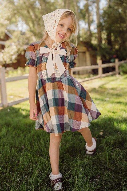 A young girl wearing the Ivy City Co Mini Alice Dress in Maize Gingham, featuring a smocked bodice and puff sleeves, smiles while standing on green grass with sunlight and trees in the background.
