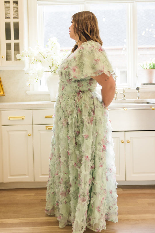 A woman wearing the Ivy City Co Florentina Dress in Pistachio Aquarelle Fleur stands in a bright kitchen, gazing out the window. White cabinets, fresh flowers, and a farmhouse sink can be seen in the background.