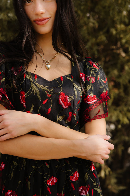 A woman stands with arms crossed, wearing Ivy City Co’s Flora Dress in Romantic Roses—a black dress adorned with embroidered red roses and a delicate black mesh overlay. She accessorizes with a gold heart necklace. Against a backdrop of softly blurred green foliage, her partially visible face holds a slight smile, embodying the elegance of the Flora Dress.