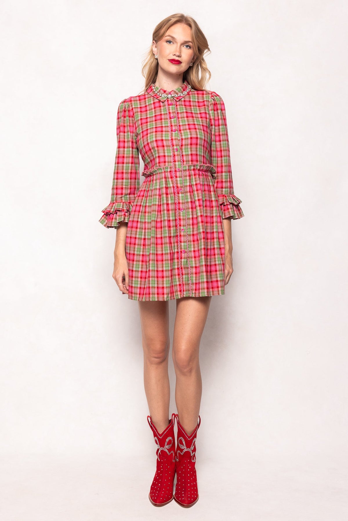 A woman with blonde hair stands against a white background, wearing a red and pink plaid dress with ruffled sleeves and collar. She completes her look with the Channing Bootie in Red by Ivy City Co, making a bold statement with these standout suede cowboy booties.