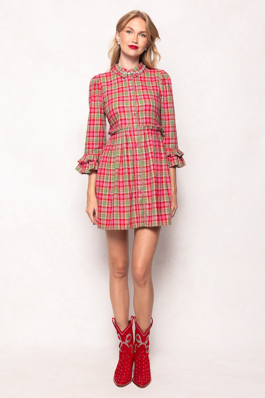 A woman with blonde hair stands against a white background, wearing a red and pink plaid dress with ruffled sleeves and collar. She completes her look with the Channing Bootie in Red by Ivy City Co, making a bold statement with these standout suede cowboy booties.