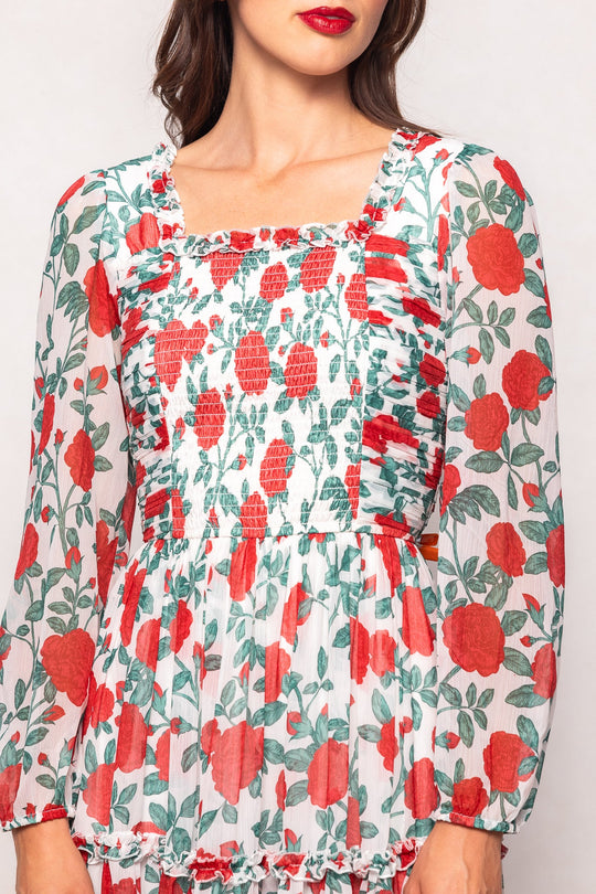 A woman wearing the Selena Dress in Red Roses by Ivy City Co—a maternity-friendly chiffon midi dress featuring long sleeves, a square neckline, a ruched bodice, and a sheer white background adorned with bright red flowers and green leaves.