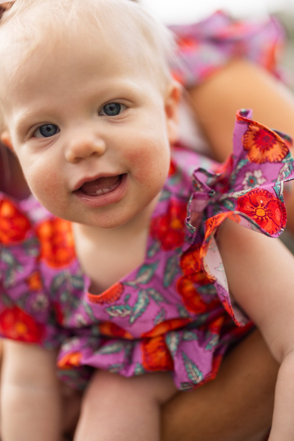 A smiling baby with blue eyes and light hair wears the Ivy City Co Baby Hattie Romper in Thistle Flower Poppy Woodblock, featuring purple floral prints and ruffled sleeves, looking directly at the camera.