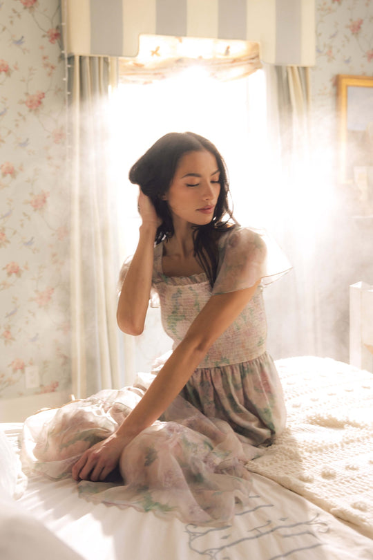 A woman wearing the Ivy City Co Rhiannon Dress in Painted Bloom sits on a bed in a softly lit room, sunlight streaming behind her and creating a dreamy atmosphere.