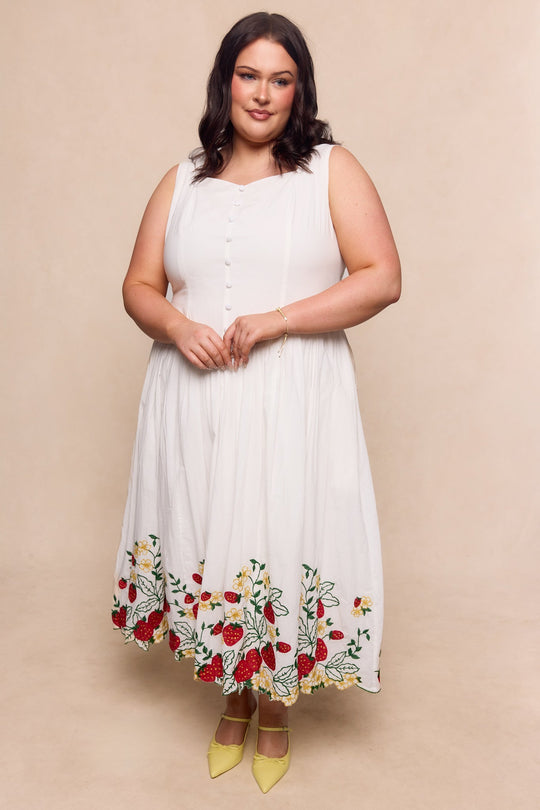 A woman stands against a beige backdrop, softly smiling as she wears the Ivy City Co Clarissa Dress in Strawberry—a sleeveless white dress featuring colorful floral embroidery near the hem—paired with yellow heels.