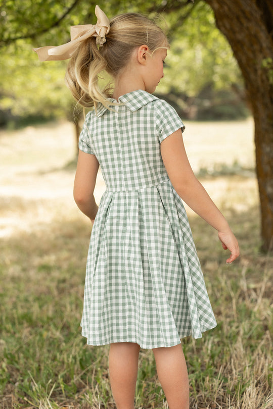 A young girl with a beige bow in her blonde hair wears the Ivy City Co Mini Amber Dress in Sage Gingham, featuring a pleated skirt, as she stands outdoors on the grass beneath a tree, facing away.