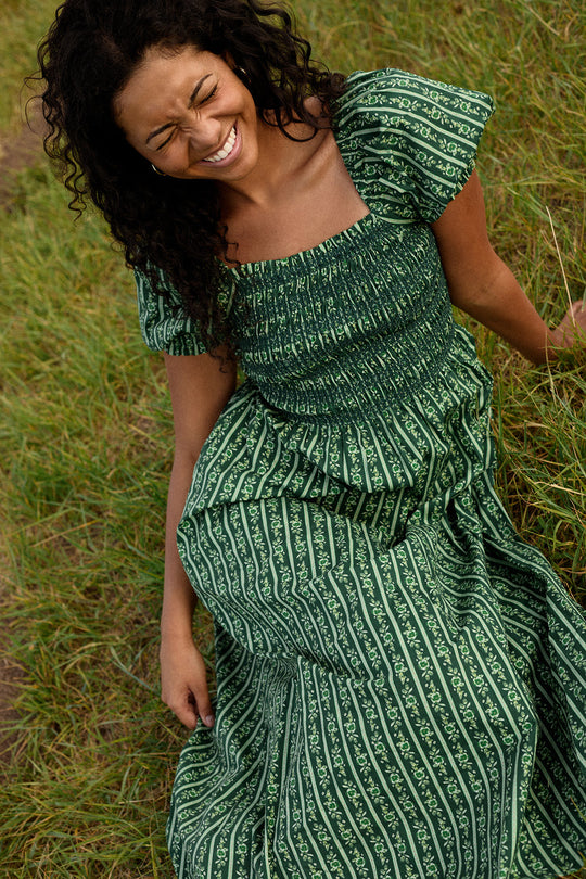 A woman with curly hair sits on the grass, smiling with closed eyes, wearing the Ivy City Co Sunday Best Dress in Pine Felicity Floral—a stylish choice featuring puff sleeves and designed to be maternity friendly.
