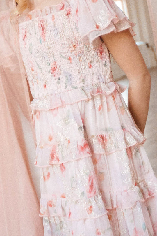 A person wearing the Ivy City Co Mini Winnie Dress in Pink Floral—featuring a light pink hue, floral print, ruffled details, a smocked bodice, and glitter accents—standing indoors.