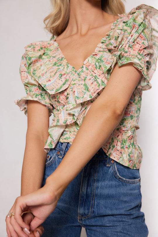 A woman wearing the Julian Top in Cream by Ivy City Co—featuring ruffles, a floral pattern, and puff sleeves—stands with her arms gently crossed in front of her body, paired with blue jeans.