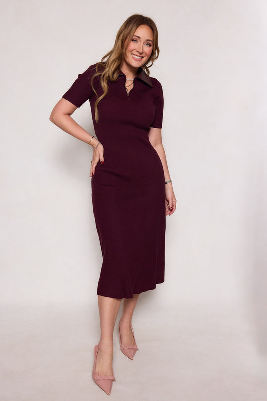 A woman stands smiling against a plain light background, wearing the Ora Dress in Burgundy by Ivy City Co—a short-sleeve midi dress with a structured collar—styled with pink heels and minimal jewelry, one hand on her hip.