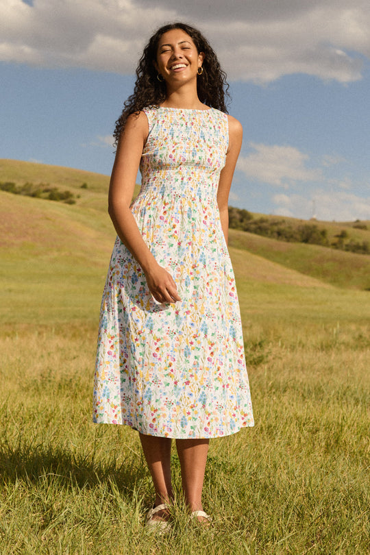 Wearing the Ana Dress in Floral Fields by Ivy City Co, a woman stands smiling in a grassy field with rolling hills and a partly cloudy sky behind her. The sleeveless midi dress features a smocked bodice and a maternity-friendly design, offering both comfort and effortless style.