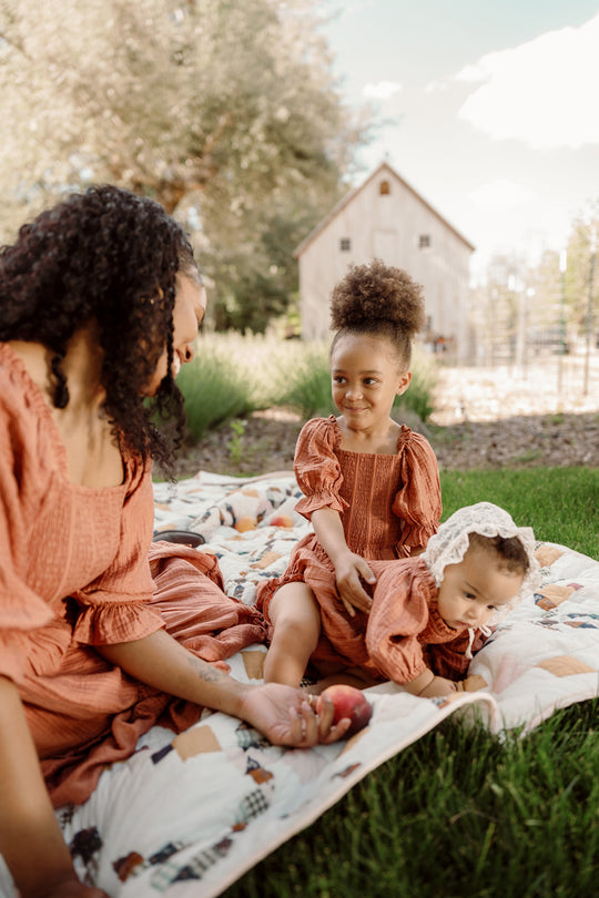 A woman and two young children in matching rust-colored outfits sit on a quilt outdoors, with a barn in the background. The woman wears the Ivy City Co Mini Madeline Dress in Apple Cider, featuring delicate puff sleeves and a smocked bodice. One child smiles at the woman, while the other crawls on the blanket.